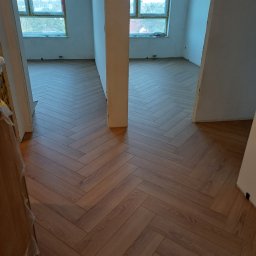 Skrzynka synka - Nowe panele ułożone we wzór jodełki francuskiej w jasnym, przestronnym wnętrzu z dużymi oknami i surowymi ścianami, widok z perspektywy wejścia do pokoju.