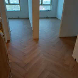 Nowe panele ułożone we wzór jodełki francuskiej w jasnym, przestronnym wnętrzu z dużymi oknami i surowymi ścianami, widok z perspektywy wejścia do pokoju.