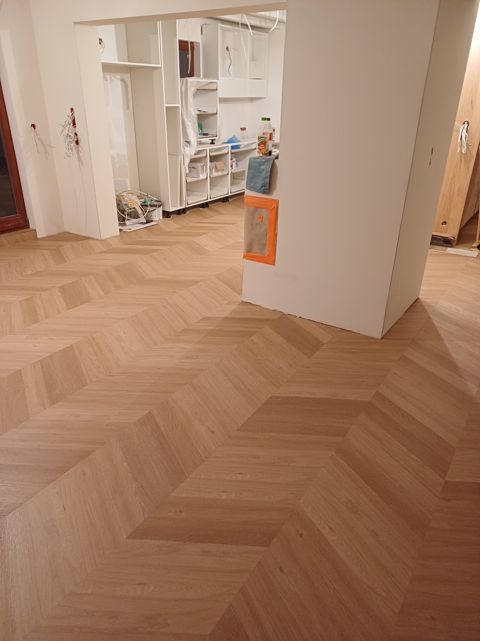Jasny parkiet ułożony we wzór jodełki francuskiej, widoczny w trakcie prac wykończeniowych wnętrza, z elementami szafy w zabudowie i przygotowaniami do instalacji elektrycznej.