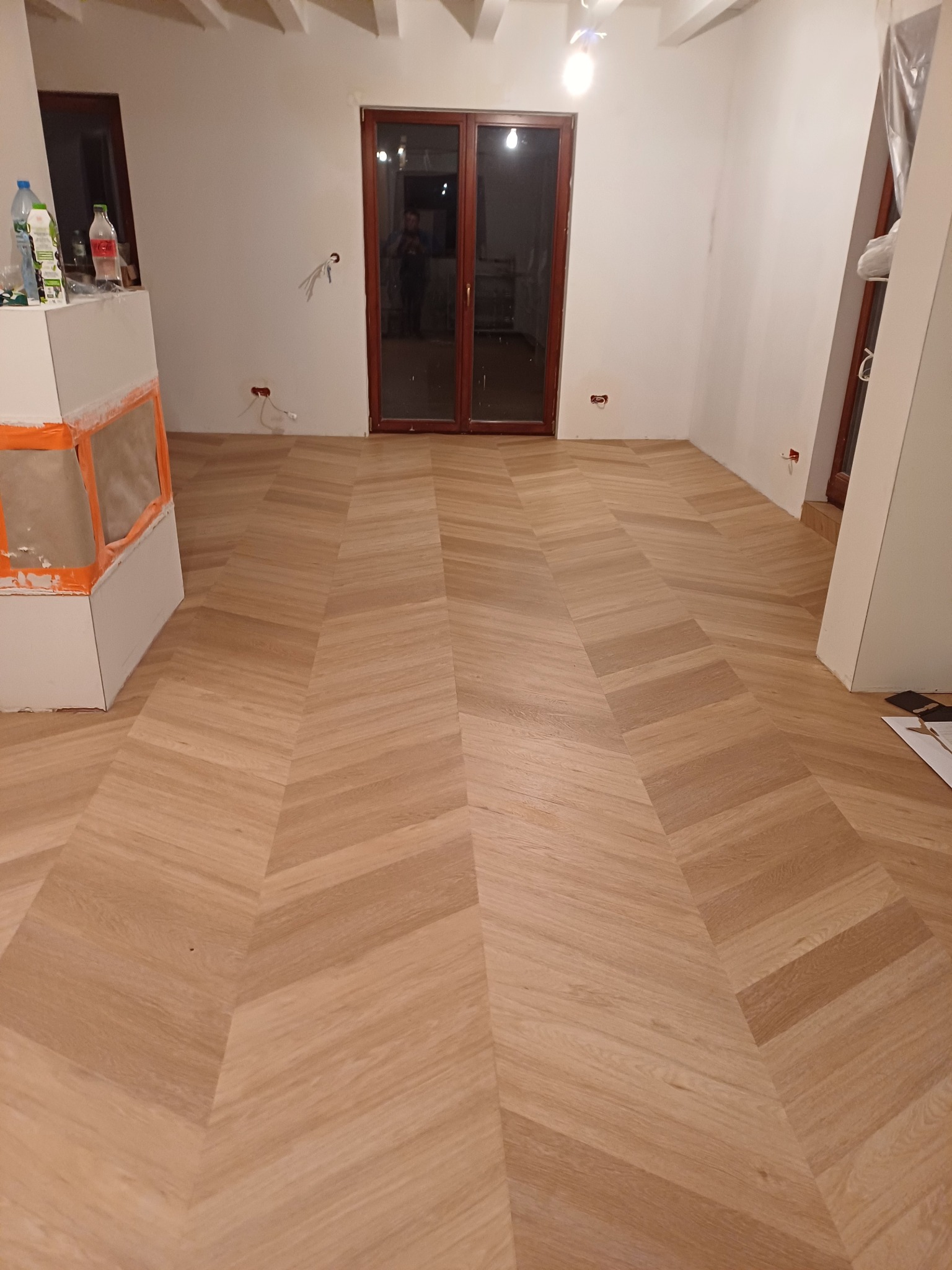 Nowo ułożona podłoga z jasnego drewna w jodełkę francuską, widoczne gniazdka elektryczne w ścianie, fragment kominka zabezpieczonego folią malarską oraz drewniane drzwi w tle.