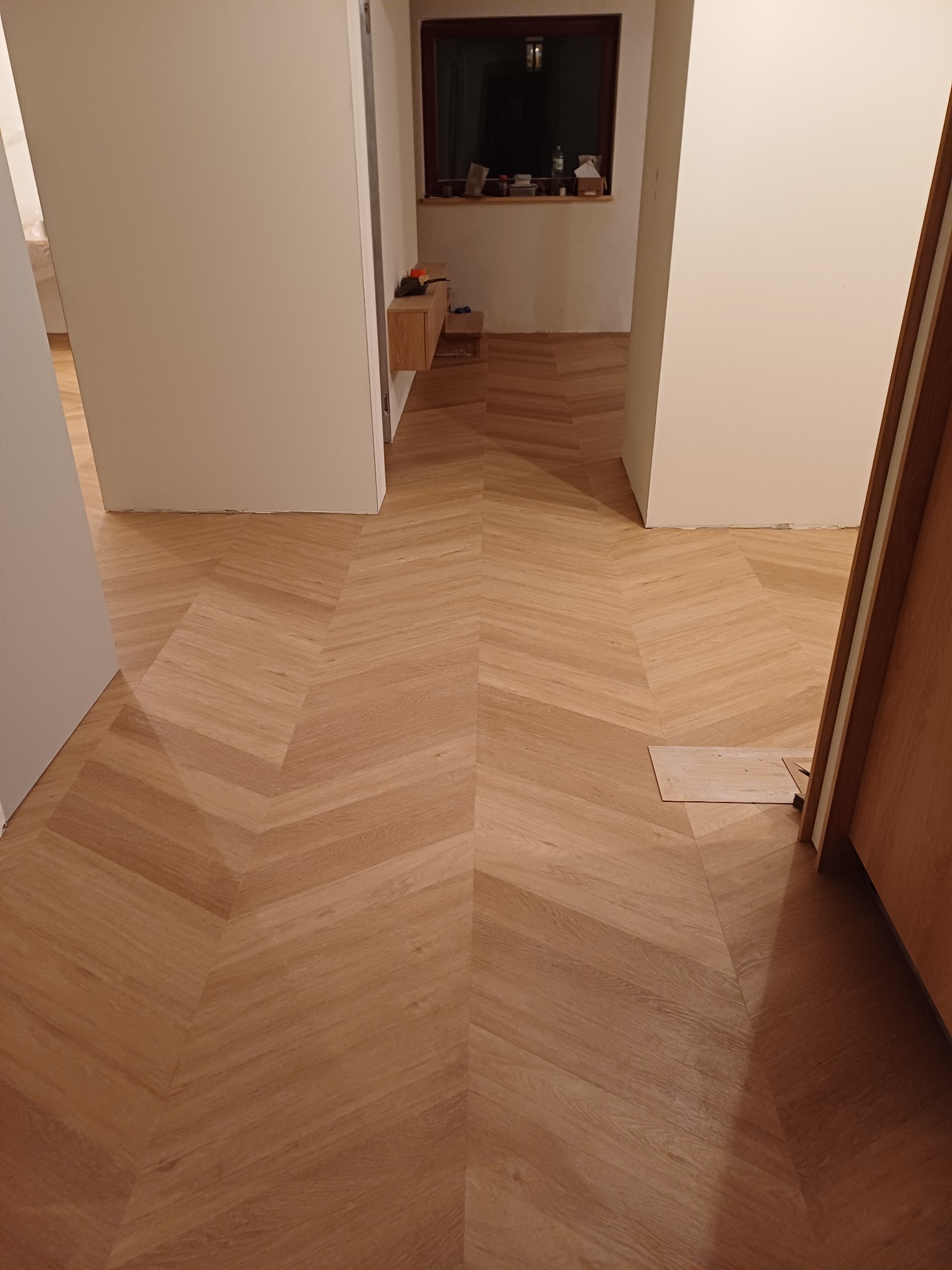 Ułożony wzór jodełki z jasnego drewna na podłodze, widoczny fragment niedokończonego montażu przy progu drzwiowym.