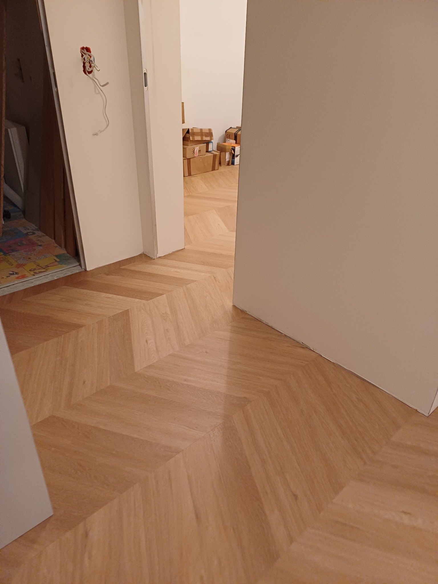 Jasny parkiet ułożony we wzór jodełki francuskiej, widok z perspektywy podłogi w kierunku otwartych drzwi, widoczne kartony w oddali.