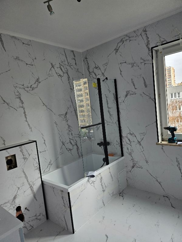 Wykończona łazienka z wanną obudowaną panelami imitującymi marmur, z czarną ramą kabiny prysznicowej i baterią, oraz z oknem, na parapecie którego leży wkrętarka.