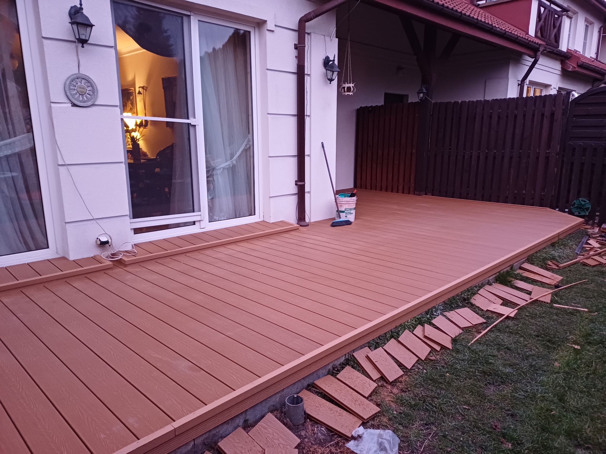 Nowo wybudowany taras z desek kompozytowych w kolorze jasnego drewna, częściowo ukończony, z widocznymi elementami montażowymi i narzędziami, przylegający do jasnego budynku z dużymi oknami.