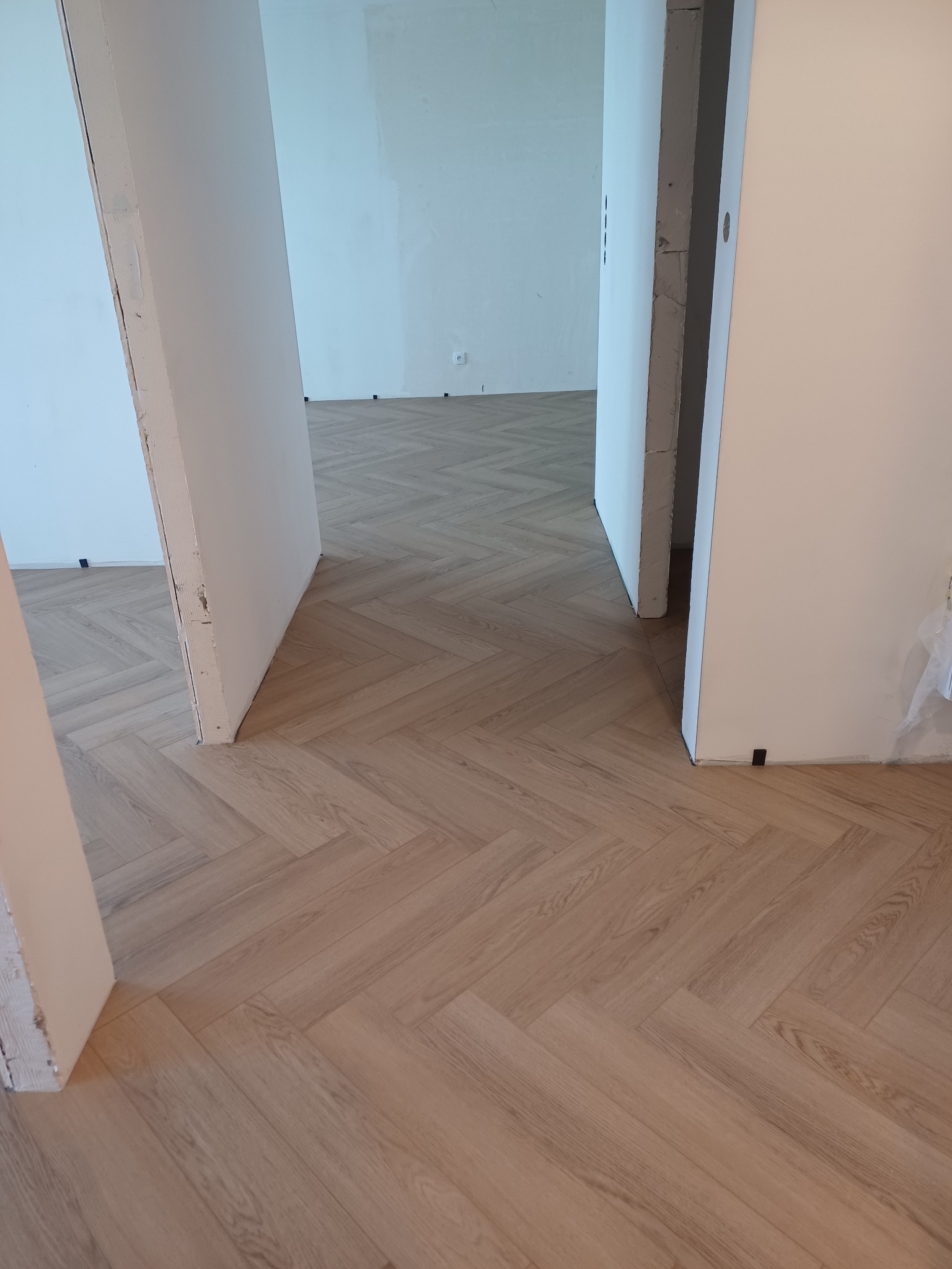 Jasny, parkiet ułożony we wzór jodełki francuskiej w nowym mieszkaniu, widoczny przez otwarte drzwi, ściany w trakcie malowania.