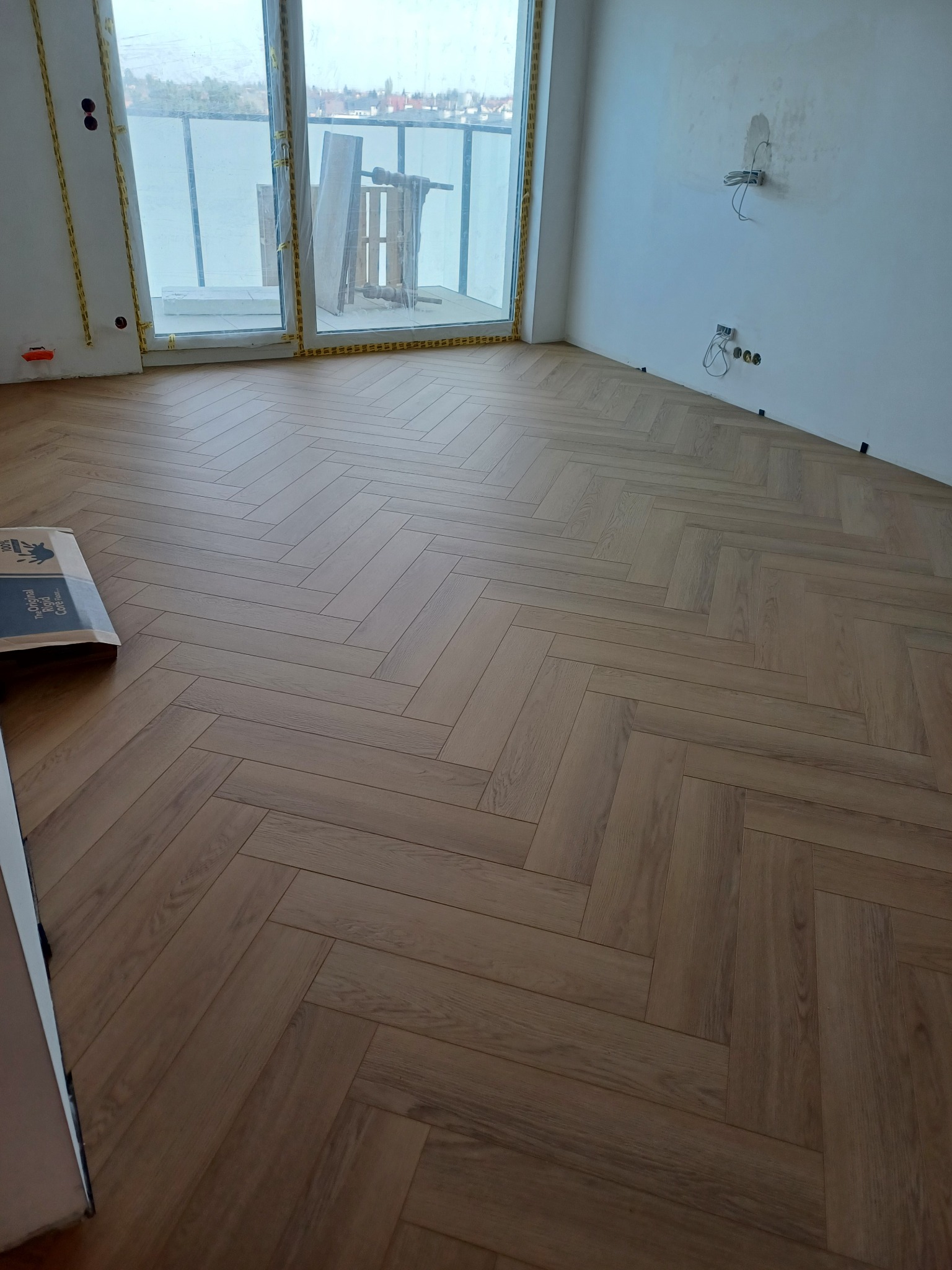 Ułożona podłoga z paneli w jodełkę w nowym mieszkaniu, widoczne paczki paneli i niezabezpieczone kable elektryczne na ścianie.