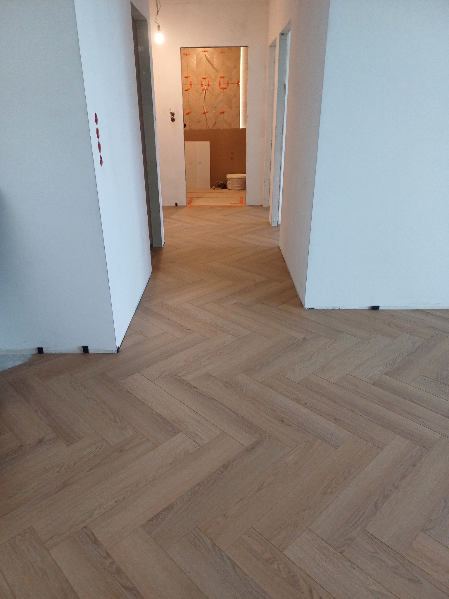 Ułożona podłoga z paneli w jodełkę w korytarzu, widoczne czarne kliny dystansowe przy ścianie, w tle prace wykończeniowe w łazience.
