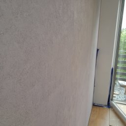 Ekskluzywne ściany - Ściana z dekoracyjną, szarą fakturą, zabezpieczona niebieską taśmą malarską przy białej ramie okiennej z widokiem na zieleń za oknem.