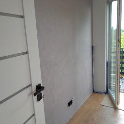 Ekskluzywne ściany - Świeżo pomalowane wnętrze z efektem dekoracyjnym na ścianie, widoczna biała stolarka drzwiowa z czarną klamką i wyeksponowane gniazdka elektryczne pod ścianą.