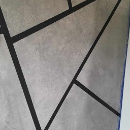 Ekskluzywne ściany - Ściana pomalowana na szaro z geometrycznym wzorem z czarnych linii, zabezpieczona niebieską taśmą malarską przy suficie i podłodze.
