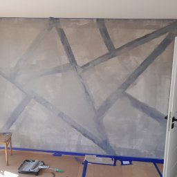 Ekskluzywne ściany - Ściana w trakcie malowania z geometrycznym wzorem wyznaczonym taśmą malarską, widoczny wałek i kuweta z farbą, zabezpieczona podłoga kartonem, krzesło z plamami farby w rogu pomieszczenia.