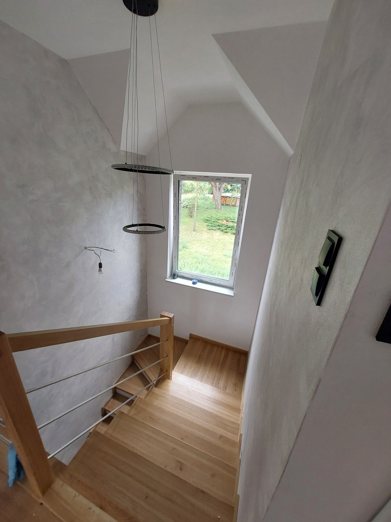 Klatka schodowa z drewnianymi schodami i metalową balustradą, ściany pokryte dekoracyjnym tynkiem w odcieniach szarości, wisząca lampa z okrągłymi elementami, widok z góry.