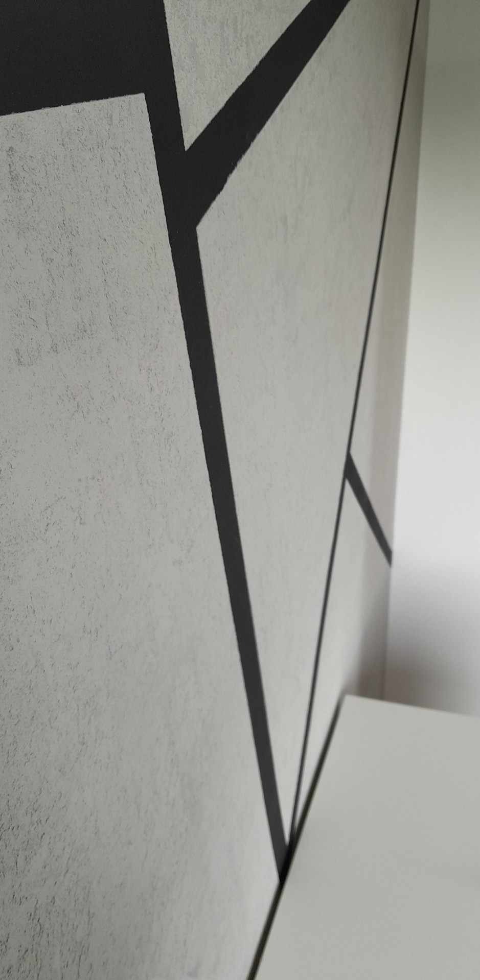 Ściana z szarą, teksturowaną farbą i geometrycznymi, czarnymi liniami, częściowo widoczny biały mebel w rogu.
