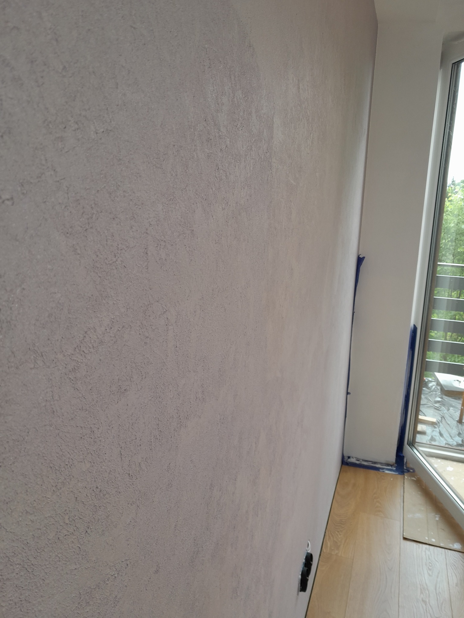 Ściana z dekoracyjną, szarą fakturą, zabezpieczona niebieską taśmą malarską przy białej ramie okiennej z widokiem na zieleń za oknem.