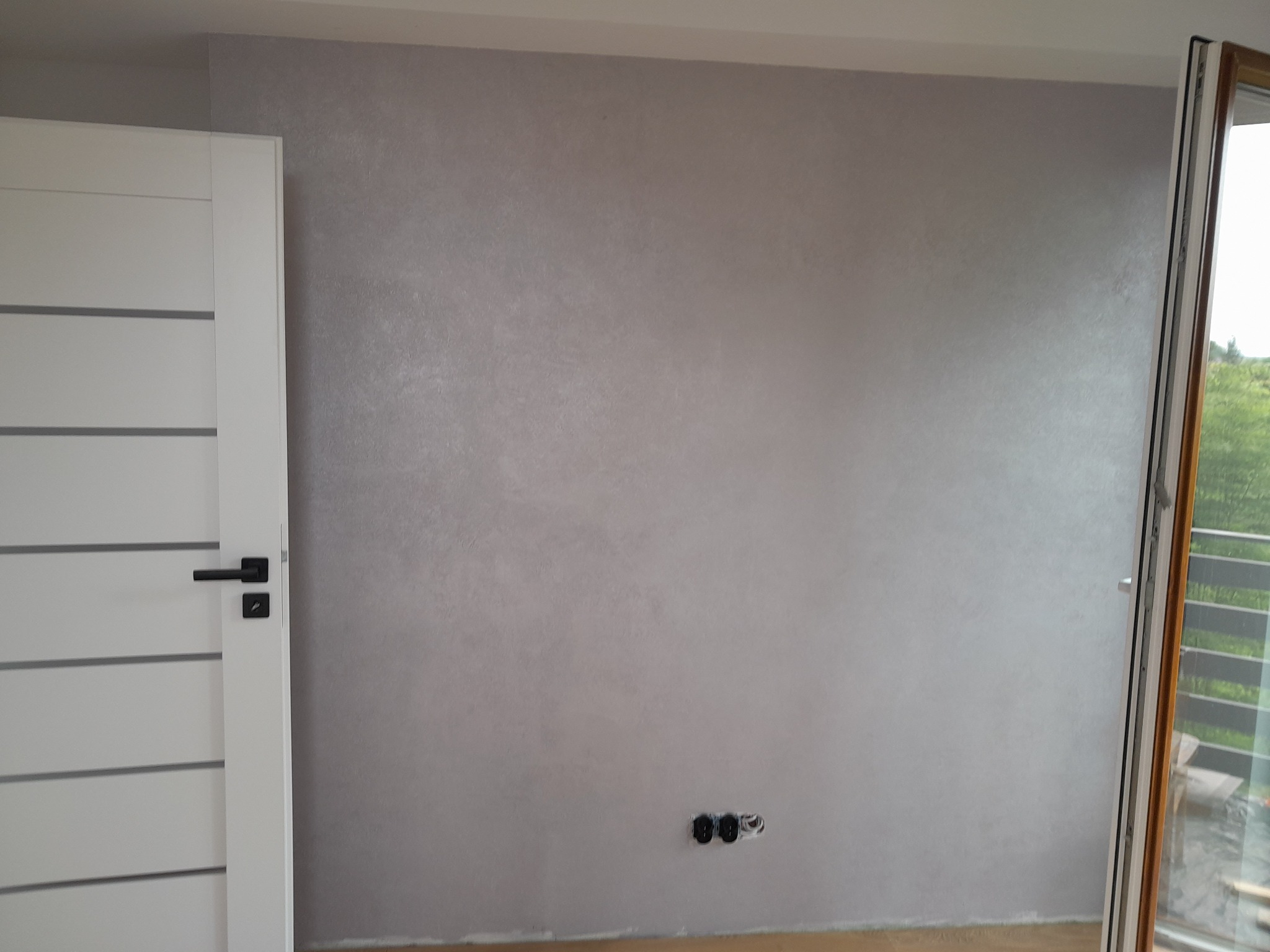Ściana pomalowana farbą dekoracyjną w odcieniach szarości z widocznymi gniazdkami elektrycznymi, obok białe drzwi z poziomymi frezami i czarną klamką oraz fragment okna balkonowego.