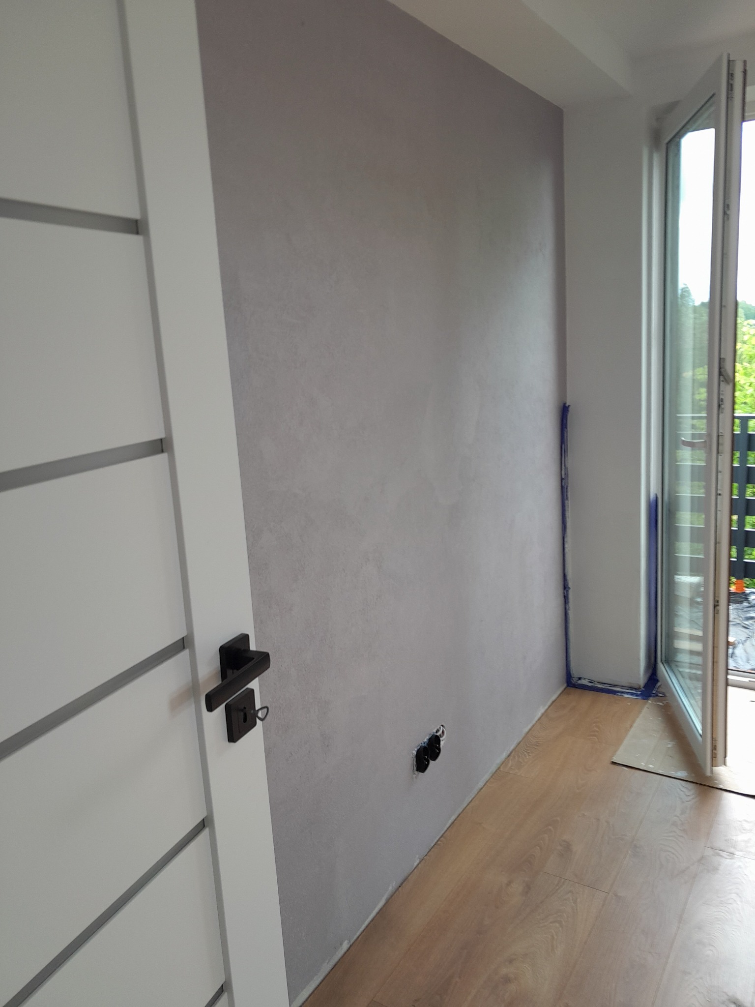 Świeżo pomalowane wnętrze z efektem dekoracyjnym na ścianie, widoczna biała stolarka drzwiowa z czarną klamką i wyeksponowane gniazdka elektryczne pod ścianą.