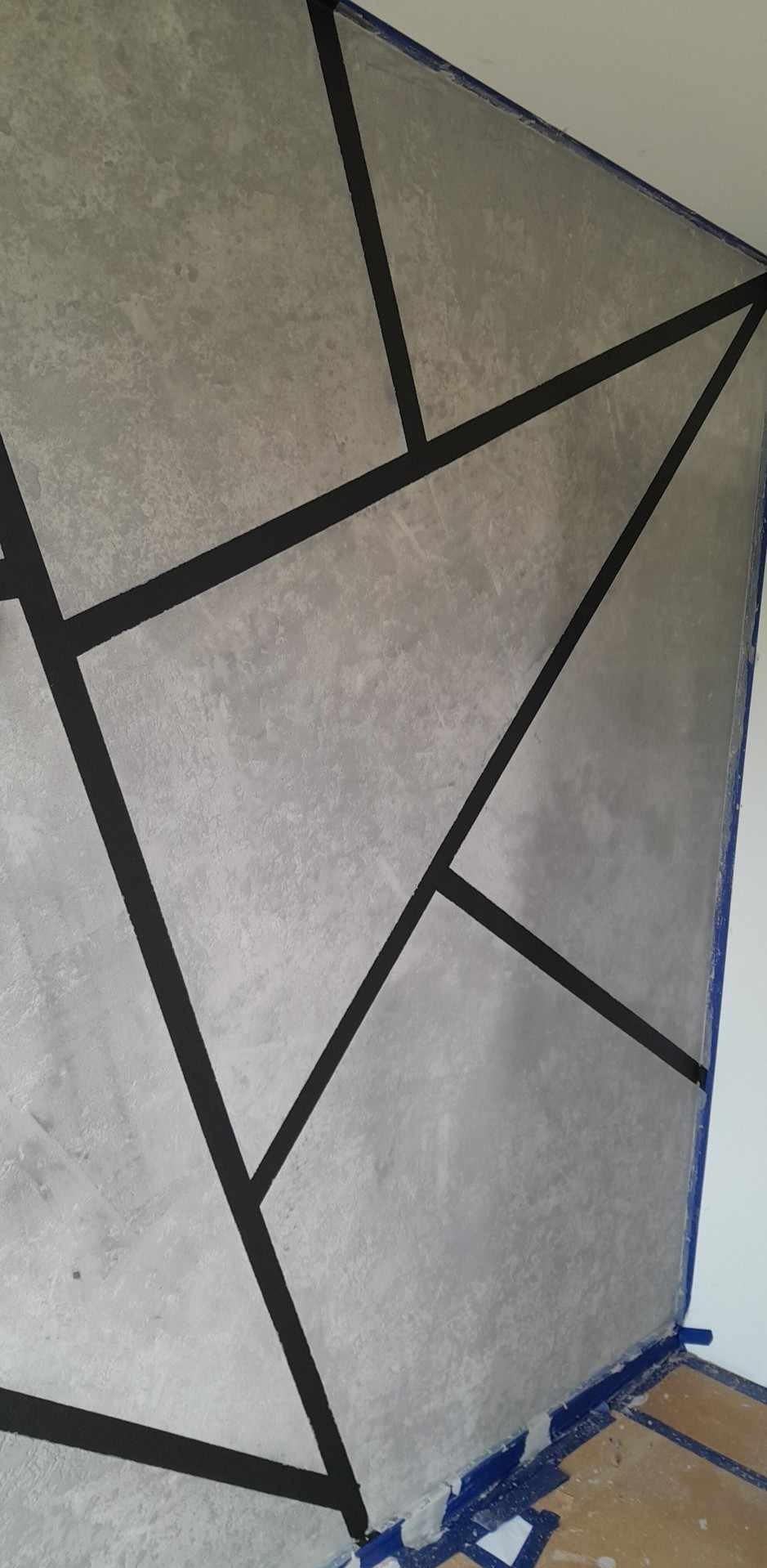 Ściana pomalowana na szaro z geometrycznym wzorem z czarnych linii, zabezpieczona niebieską taśmą malarską przy suficie i podłodze.