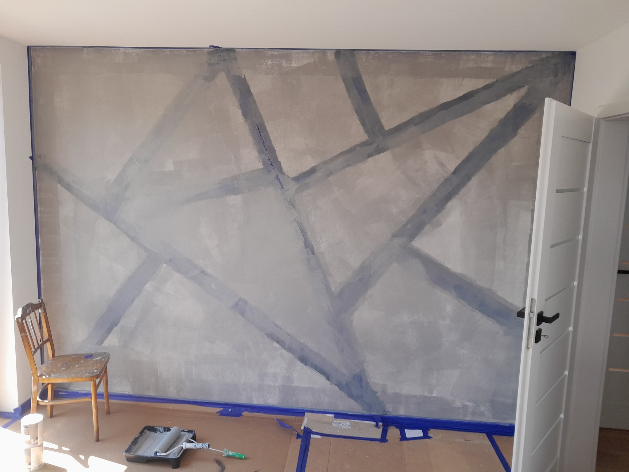 Ściana w trakcie malowania z geometrycznym wzorem wyznaczonym taśmą malarską, widoczny wałek i kuweta z farbą, zabezpieczona podłoga kartonem, krzesło z plamami farby w rogu pomieszczenia.
