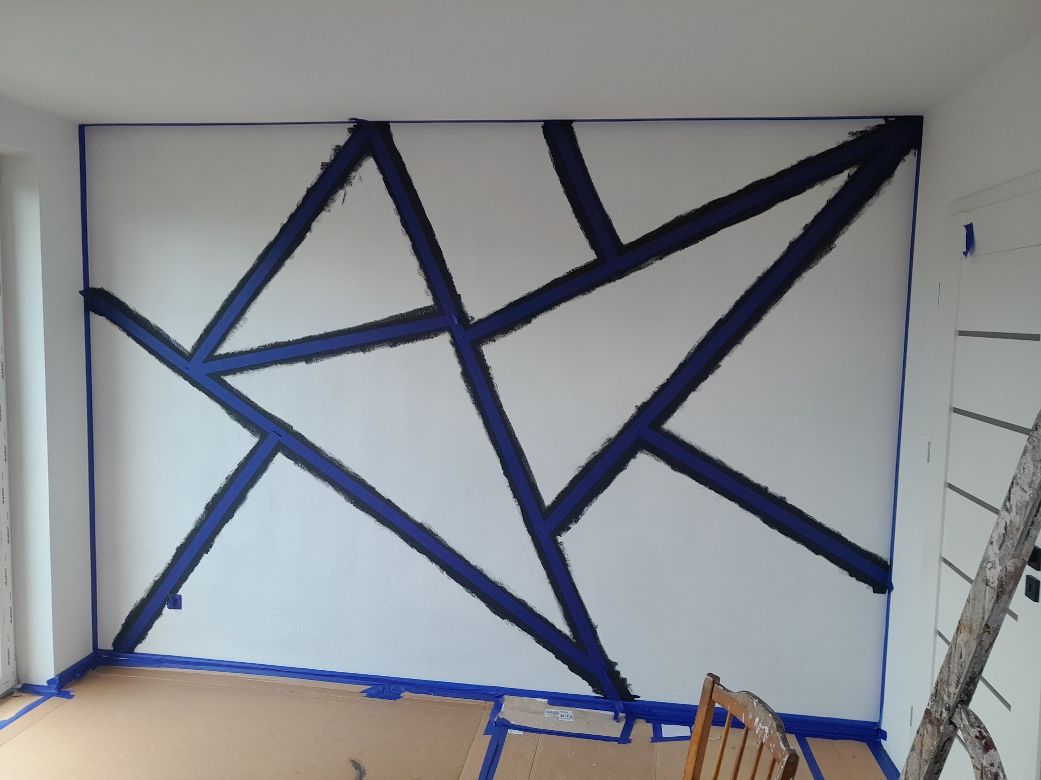 Ściana z geometrycznym wzorem malowanym na biało, zabezpieczona niebieską taśmą malarską, w trakcie tworzenia.