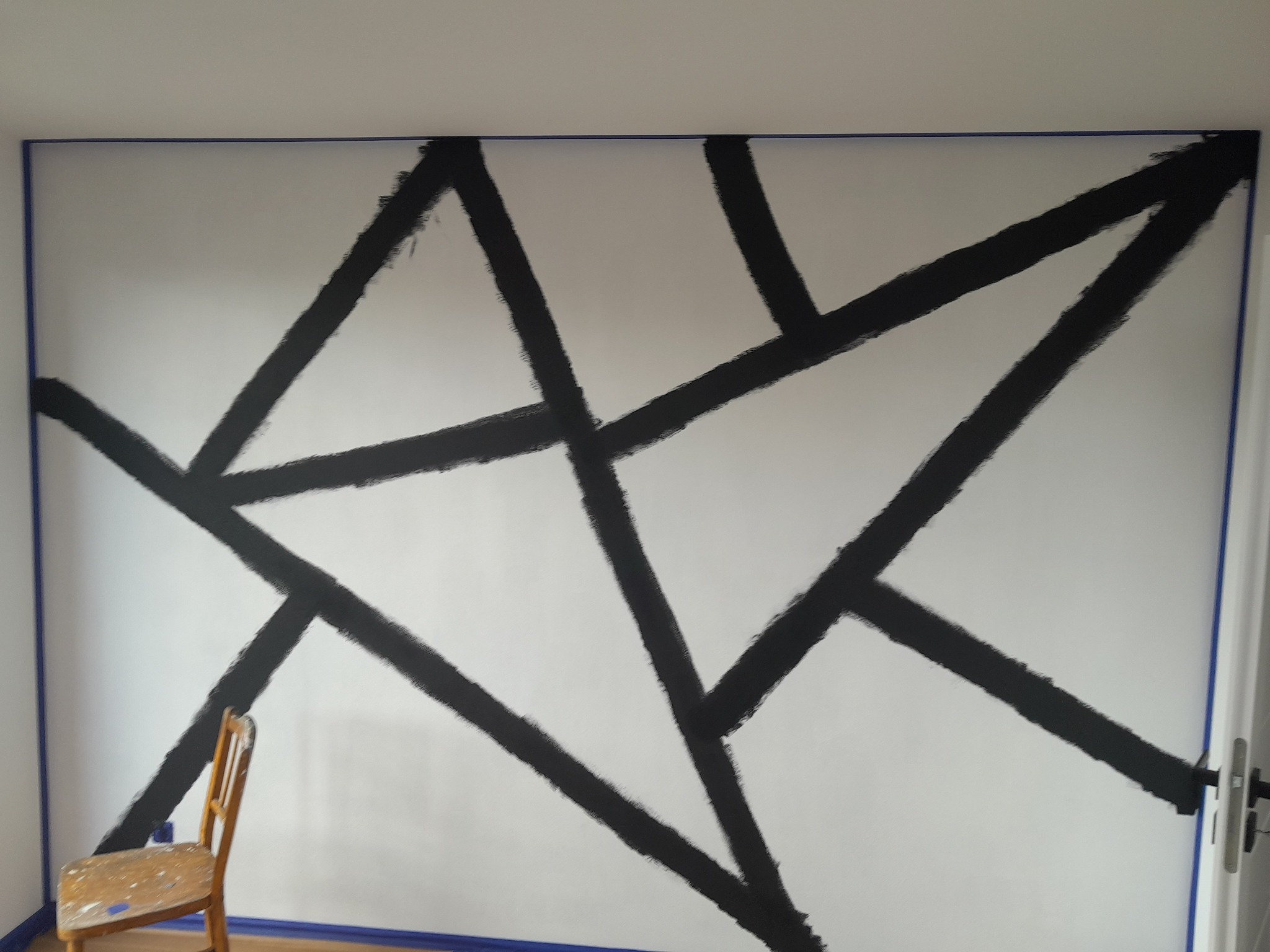 Ściana z geometrycznym wzorem z czarnych pasów, malowana w Katowicach, zabezpieczona taśmą malarską, z widocznym krzesłem malarskim.