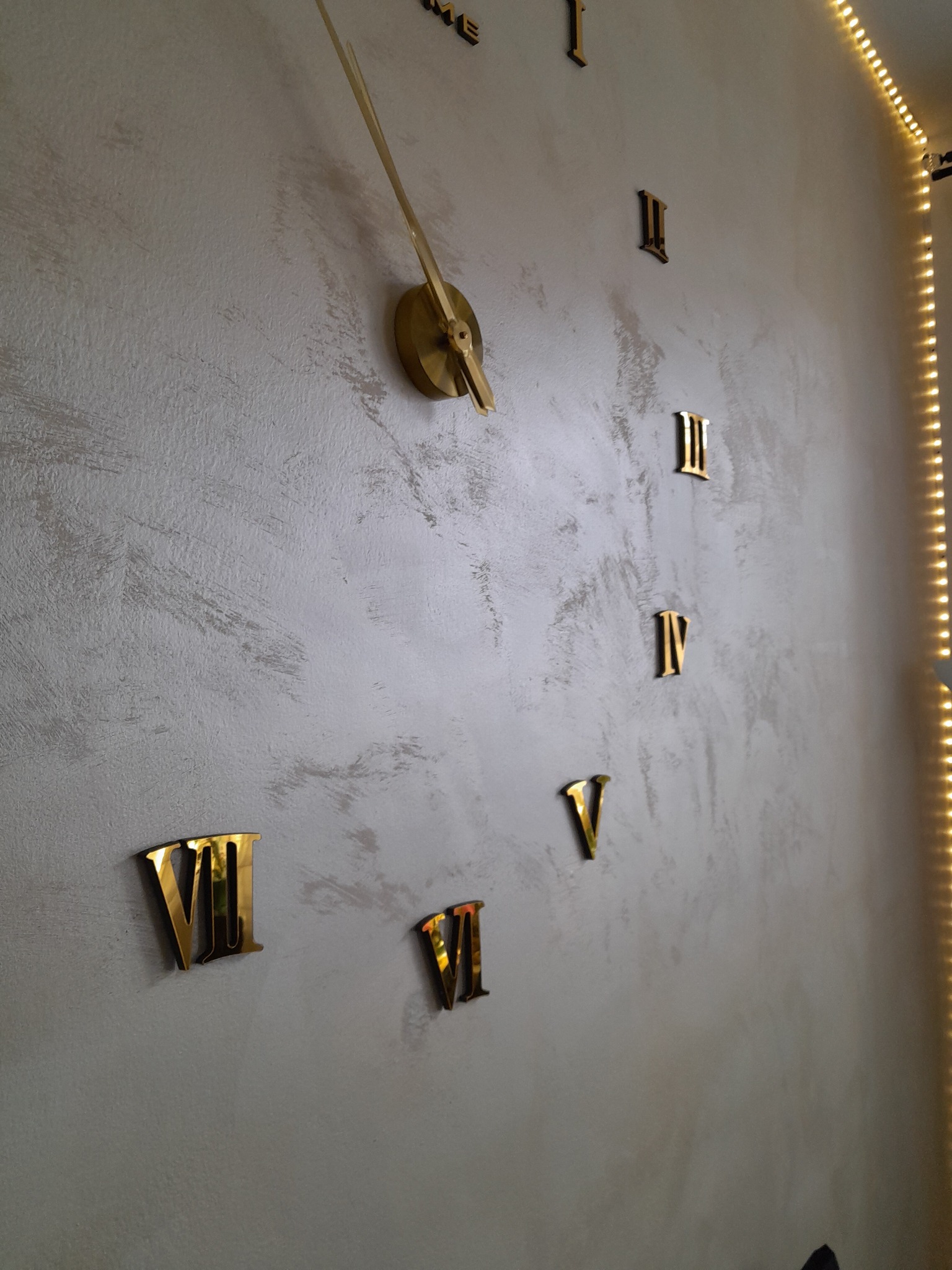 Dekoracyjna ściana z nałożonym tynkiem strukturalnym i dużym zegarem z złotymi rzymskimi cyframi, oświetlona taśmą LED.
