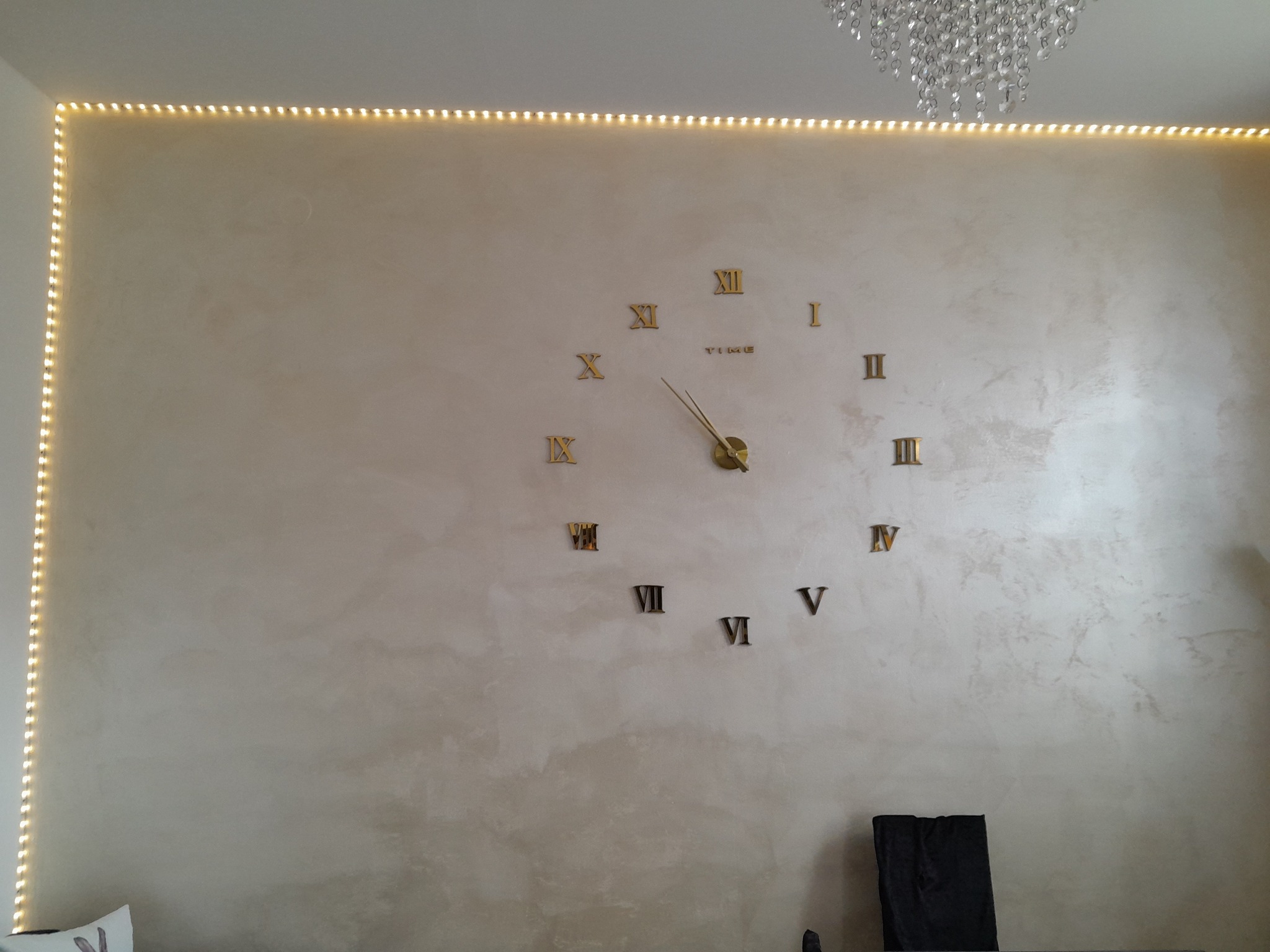 Ściana z dekoracyjnym tynkiem w odcieniach beżu, oświetlona listwą LED, z naklejonym złotym zegarem rzymskim i kryształowym żyrandolem w tle.