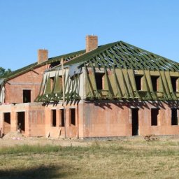 Dachiman - Surowy stan domu z cegły z widoczną konstrukcją dachu i oknami, w tle zieleń i błękitne niebo, dwóch mężczyzn przy budynku.