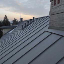 Dachiman - Szary, metalowy dach z kominem i kilkoma odpowietrznikami w Lublinie, widziany pod kątem w pochmurny dzień.