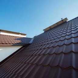 Dachiman - Brązowy dach z blachy trapezowej z widocznymi rynnami i kominem na tle błękitnego nieba.