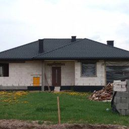 Dachiman - Dom jednorodzinny w trakcie budowy, z widocznym czarnym dachem z blachodachówki, kominami i częściowo zamurowanymi oknami, na tle zielonego trawnika z kwitnącymi mniszkami.