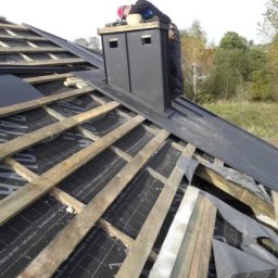 Dachiman - Pracownik w kasku montuje obróbkę blacharską komina na dachu z widoczną membraną i drewnianą konstrukcją