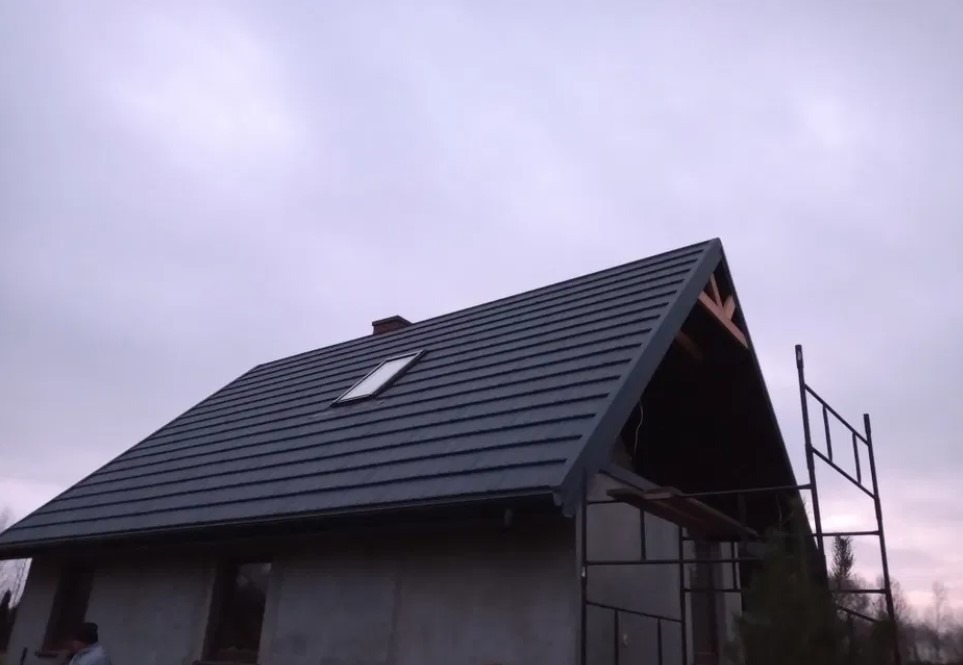 Dom w trakcie budowy z zamontowanym, grafitowym pokryciem dachowym z oknem dachowym i widoczną więźbą dachową od strony szczytu.