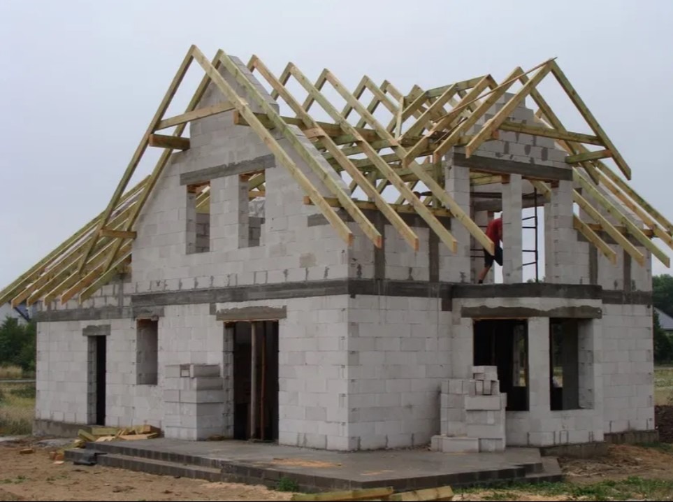 Surowy stan domu z białego bloczka, z widoczną drewnianą konstrukcją dachu w trakcie budowy i pracownikiem na drabinie wewnątrz budynku.