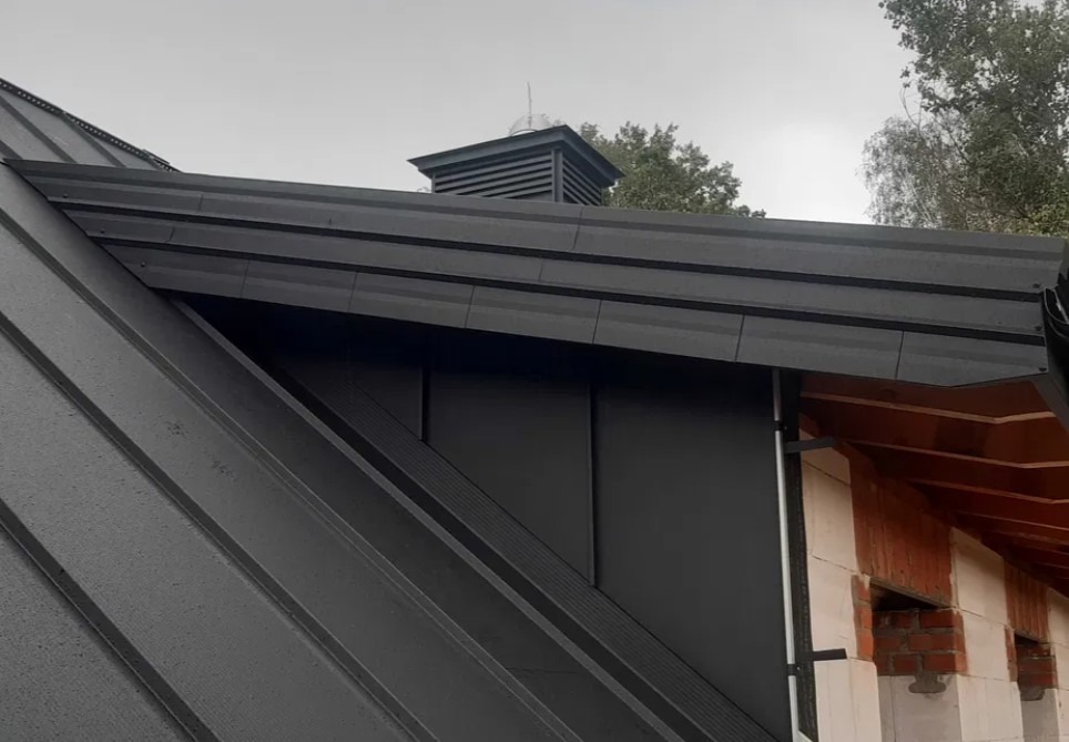 Nowoczesne, grafitowe pokrycie dachowe z blachy na skośnym dachu, widoczny kominek wentylacyjny i fragment muru budynku.