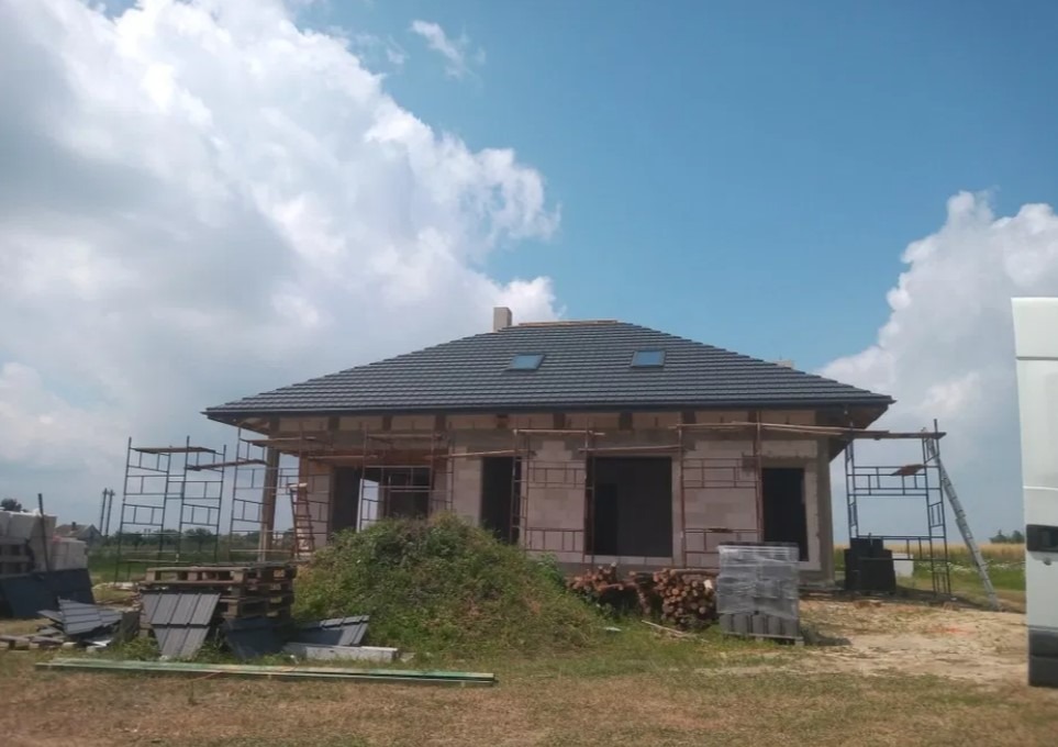 Dom jednorodzinny w trakcie budowy z zamontowaną, grafitową dachówką ceramiczną i oknami dachowymi, widoczne rusztowania i materiały budowlane na działce.