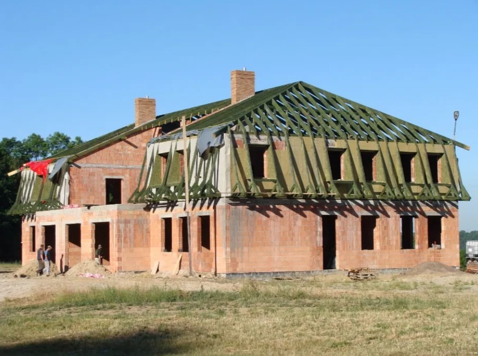 Surowy stan domu z cegły z widoczną konstrukcją dachu i oknami, w tle zieleń i błękitne niebo, dwóch mężczyzn przy budynku.