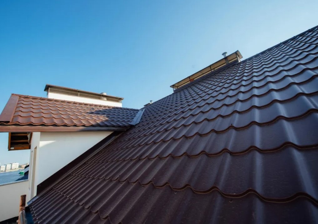 Brązowy dach z blachy trapezowej z widocznymi rynnami i kominem na tle błękitnego nieba.