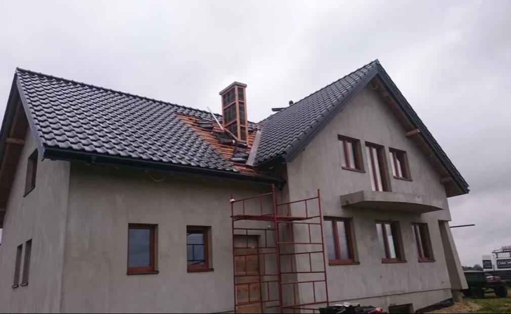 Dom w trakcie budowy z częściowo położonym, ciemnym pokryciem dachowym i widocznym kominem w trakcie wznoszenia.