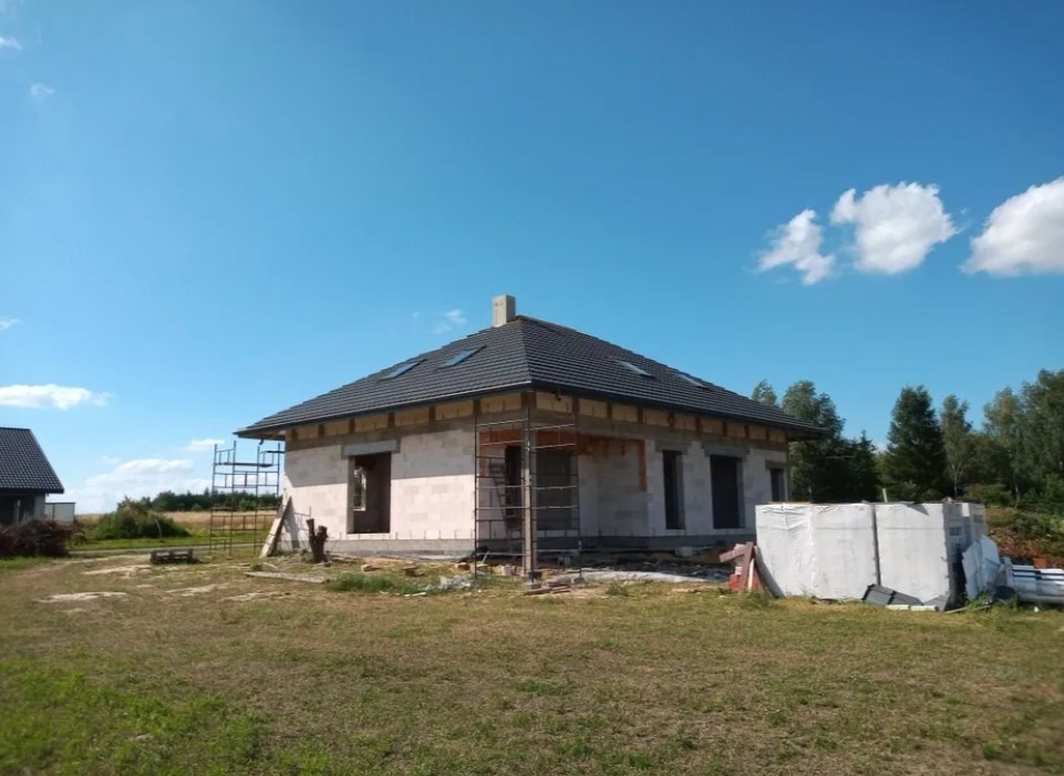 Dom jednorodzinny w trakcie budowy, widoczna konstrukcja z bloczków, czarna dachówka na dachu z oknami dachowymi, rusztowania przy ścianach, błękitne niebo.