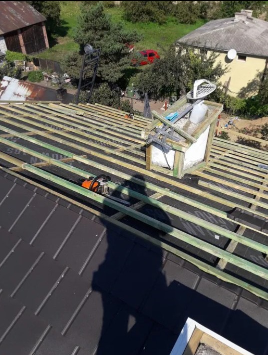 Montaż łat na dachu z czarnej blachy, widoczny komin z narzędziami i cień osoby wykonującej prace, w tle budynki i zieleń.