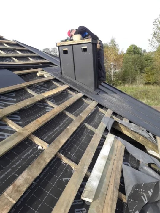 Pracownik w kasku montuje obróbkę blacharską komina na dachu z widoczną membraną i drewnianą konstrukcją