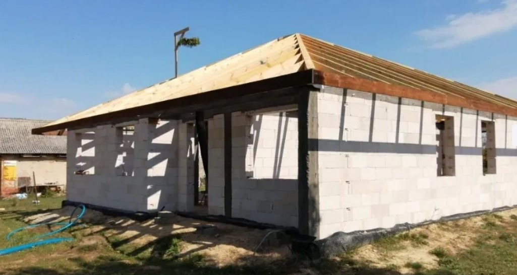 Budowa domu jednorodzinnego w trakcie wznoszenia drewnianej konstrukcji dachu, widoczne ściany z betonu komórkowego i przygotowane otwory okienne.