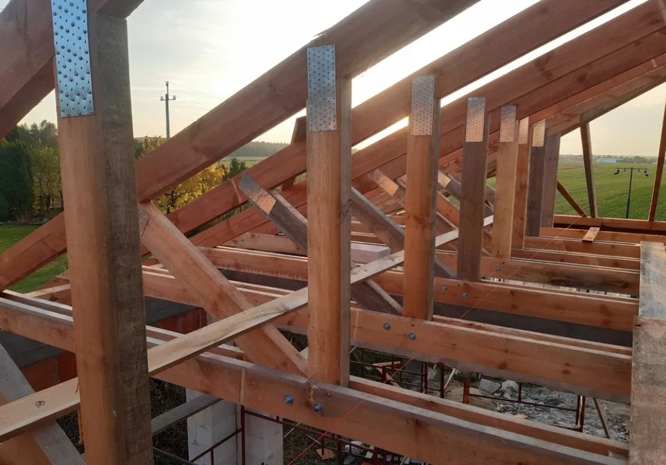 Konstrukcja drewniana dachu w trakcie budowy, widoczne elementy więźby dachowej z metalowymi łącznikami, w tle krajobraz z polami i drzewami w promieniach zachodzącego słońca.