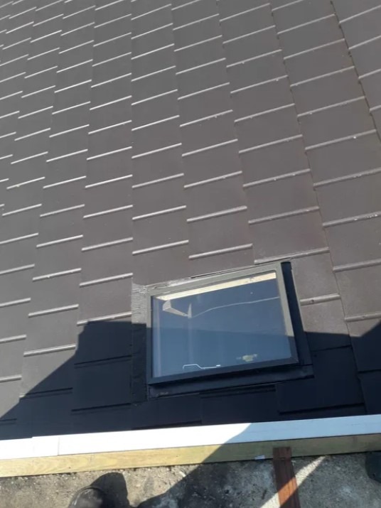 Ciemna dachówka ceramiczna z zamontowanym oknem dachowym, widok z góry na fragment dachu z rynną i fragmentem buta w rogu kadru.