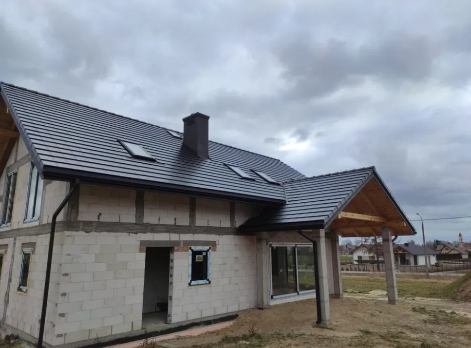 Dom w trakcie budowy z nowym, ciemnoszarym pokryciem dachowym z dachówki ceramicznej, widoczne okna dachowe i czarny komin, zamontowane rynny, ściany z białego bloczka.