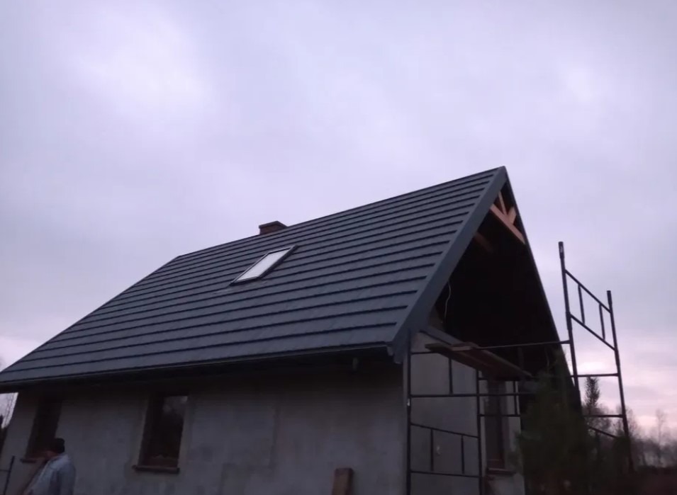 Dom z nowym, grafitowym dachem z blachy trapezowej, widoczne okno dachowe, komin i fragment niezadaszonego wejścia z elementami więźby dachowej, obok rusztowanie.