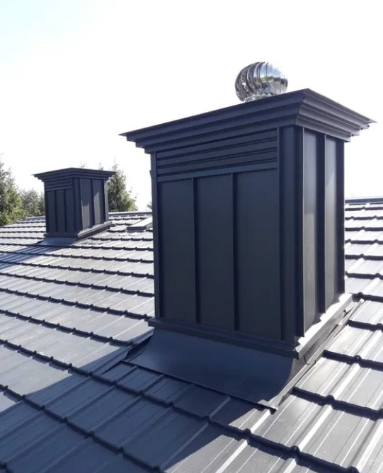 Metalowy dach z dwoma kominami w nowoczesnym stylu, jeden z turbiną wiatrową, na tle jasnego nieba i zieleni drzew.