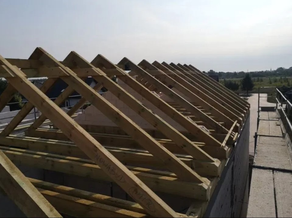 Drewniana konstrukcja więźby dachowej w trakcie montażu, widoczna z boku budynku, z rusztowaniem w tle i napisami na elementach konstrukcyjnych.