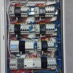 Otwarta szafa elektryczna z zainstalowanymi bezpiecznikami, przewodami w kolorach czerwonym, niebieskim i żółto-zielonym, oraz licznikiem energii z wyświetlaczem LCD.