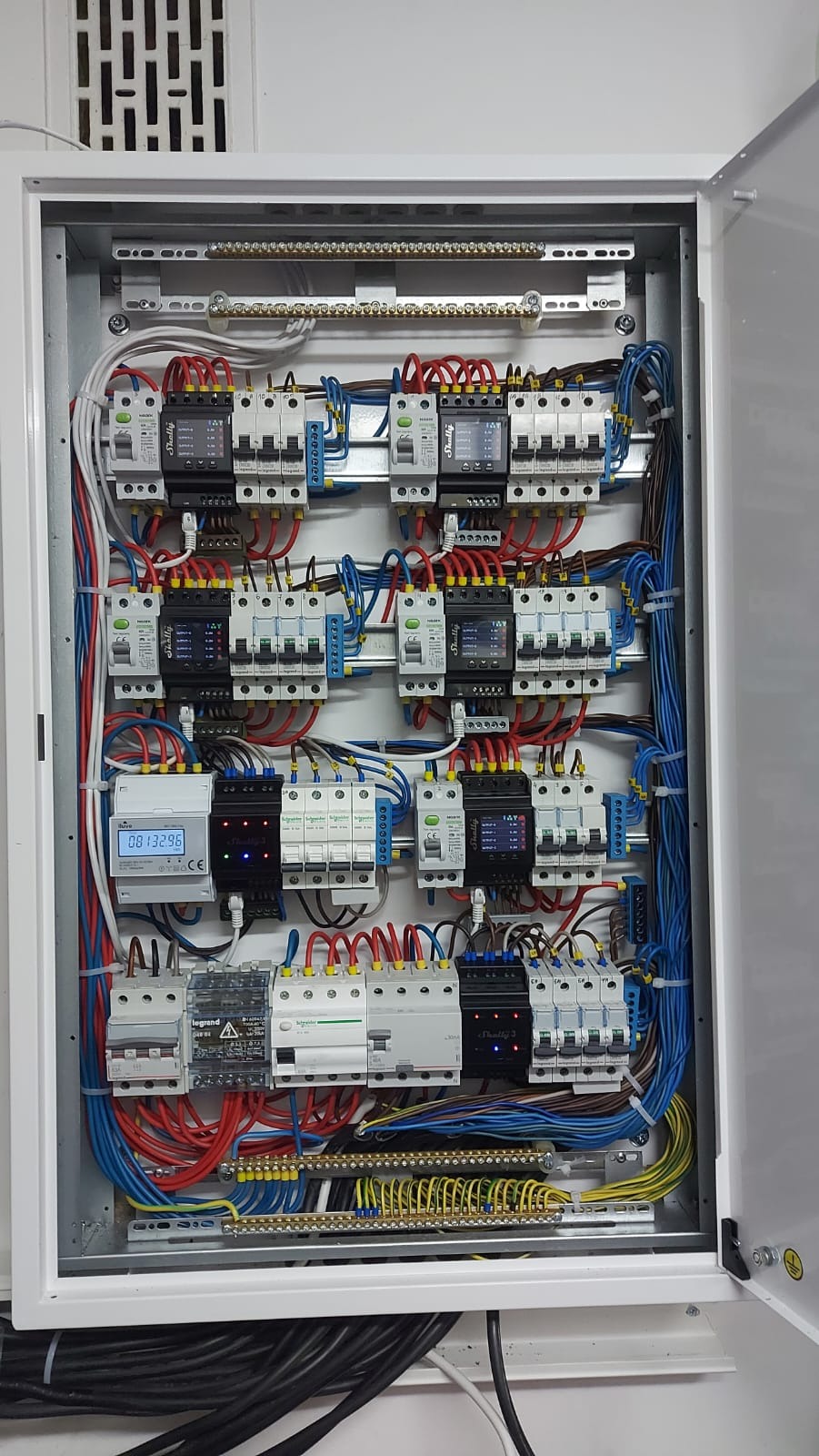 Otwarta szafa elektryczna z zainstalowanymi bezpiecznikami, przewodami w kolorach czerwonym, niebieskim i żółto-zielonym, oraz licznikiem energii z wyświetlaczem LCD.